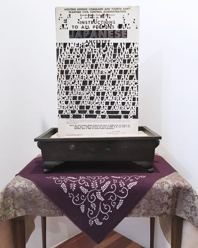 Artwork photo of a poster standing in a low, square ceramic vase atop a cloth-covered small table.